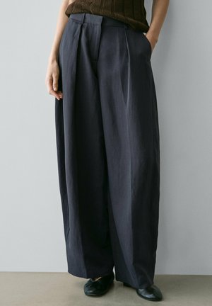 Person trägt lockere, dunkel gefärbte Hose mit weitem Bein, schwarze Schuhe und einen braunen Strickpullover, steht mit einer Hand in der Tasche.