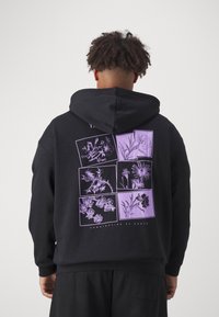 Mann trägt einen schwarzen Hoodie mit lila botanischen Skizzen, die in Rechtecken angeordnet sind, und dem Text "Kombination der Wurzeln" auf dem Rücken.