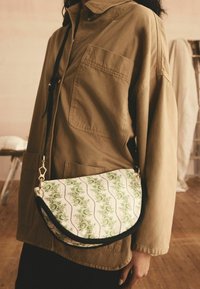 Borsa in stile hobo realizzata in un tessuto bianco con motivi floreali verdi, rifiniture nere e una tracolla in tonalità oro.