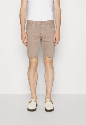 Mand iført beige denimshorts, hvid ærmeløs top og hvide slip-on sko med brune remme står mod en ensfarvet baggrund.