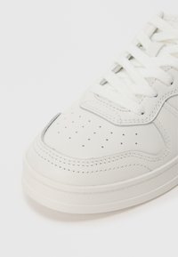 Baskets en cuir blanc avec un bout perforé, une semelle épaisse et des coutures contrastées. Présente des lacets blancs plats et une texture lisse.