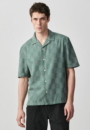 Young man wearing a short-sleeve green checkered button-up shirt and black pants, standing against a plain gray background.