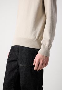 Pulôver de malha bege claro com punhos e barra canelados, combinado com calças de ganga escuras com costuras visíveis e bolsos.