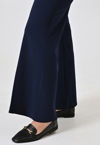 Pantaloni a gamba larga blu navy realizzati in tessuto liscio, abbinati a mocassini neri con dettaglio in metallo dorato sulla parte superiore e punta arrotondata.