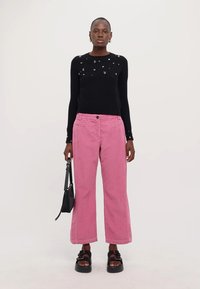 Maglione nero a maglia con decorazioni, pantaloni a gamba larga in corduroy rosa e sandali platform neri. La modella tiene una piccola borsa nera.