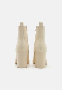 Bottines beige en similicuir avec une texture lisse, talon carré et fermeture éclair arrière. Présentent un détail de coutures verticales sur les côtés.