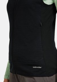 Närbild av en person som bär en svart zip-up väst med "icebreaker"-logotypen på nedre sidan, över en ljusgrön långärmad tröja.
