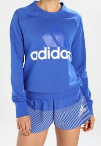 Sudadera azul con logotipo "adidas" blanco; combinada con pantalones cortos a rayas azules. La sudadera tiene un cuello redondo y puños de canalé; los pantalones cortos cuentan con cinturilla elástica.
