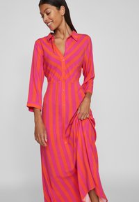 Rochie lungă, cu nasturi, cu guler, în dungi alternate roz și portocalii. Prezintă mâneci lungi și o fustă fluidă. Textură de țesătură netedă.