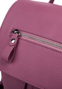 Sac à dos en cuir violet avec une finition texturée, une poche zippée à l'avant avec des accessoires en métal et un rabat arrondi.