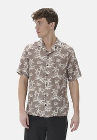 Camisa de manga corta, con botones, en tejido marrón claro con patrones de hojas blancas. Cuenta con un collar tipo camp y un bolsillo en el pecho.