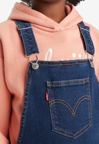 Džínové lacláče s nastavitelnými modrými popruhy a stříbrným kováním. Obsahují přední kapsu s kontrastním stehem a červenou nášivkou Levi's.