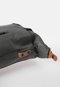 Grijze waterdichte tas met een gestructureerd oppervlak, voorzien van bruine leren accenten en oogjes. Beveiligde klep met contrasterende stikseldetails.