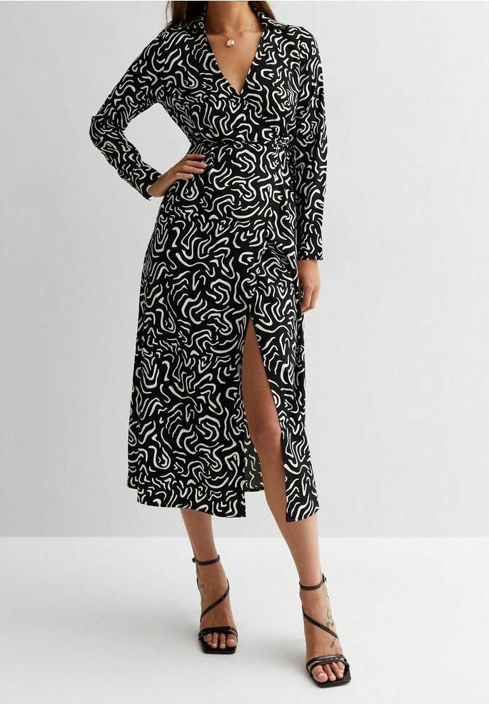 Femme portant une robe portefeuille à motifs abstraits noir et blanc, à manches longues et fente haute à la cuisse, associée à des talons noirs à brides.