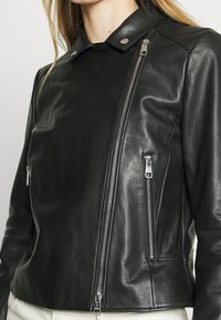 Svart läder motorcykeljacka med asymmetrisk dragkedja, tryckknappshals och två sidofickor med dragkedja. Slät textur med metalliska detaljer.