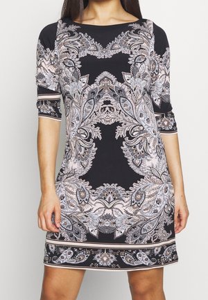 Femme portant une robe noire avec des motifs floraux et cachemire symétriques complexes beige et blanc, manches au coude et encolure ronde.
