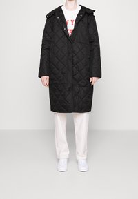 Abrigo largo acolchado negro con capucha, cierre frontal de botones y bolsillos laterales, usado sobre una camiseta gráfica blanca y pantalones claros.