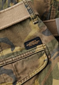 Tela con patrón de camuflaje en tonos tierra, con una cinturilla texturizada marrón. Presenta una etiqueta negra con texto naranja. Incluye detalle de bolsillo trasero.
