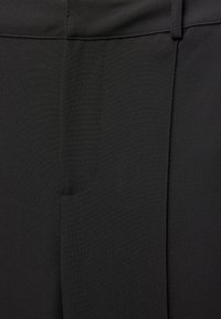 Gros plan sur un pantalon en tissu noir montrant la ceinture, les passants de ceinture et les détails de la couture centrale.