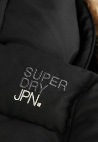 Melna pūkaina jaka ar izšūtu tekstu "SUPER DRY JPN." pelēkā un baltā krāsā, izceļot gludu, teksturētu auduma virsmu.