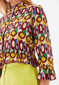 Mønstret bluse i mange farver med ståkrave, flagrende kant og puffede ærmer. Mønsteret har geometriske former i rød, grøn, blå og beige.