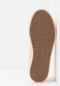 Semelle unique de basket rose clair avec un motif de bande de roulement géométrique, posée contre une surface blanche.