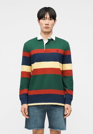 Jeune homme portant un maillot de rugby à manches longues à rayures vertes, rouges, bleu marine et jaunes, associé à un short en denim bleu.