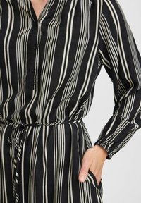 Schwarz-weiß gestreiftes Kleid mit Knopfleiste, langen Ärmeln und gebundenem Taillenbereich. Es hat eine lockere Passform und seitliche Taschen.