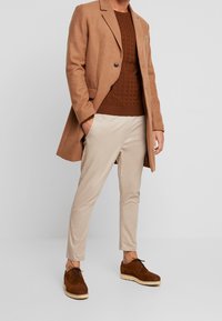 Manteau en laine marron, pull en maille marron, chinos beiges et chaussures en daim marron à semelle épaisse. Ensemble avec un design en couches et des lignes épurées.