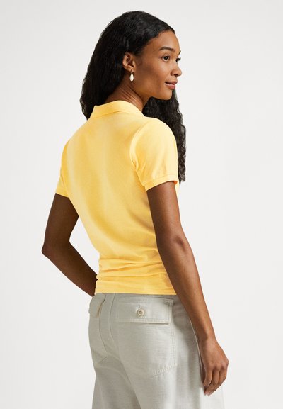 Žltá polokošeľa s krátkym rukávom, klasickým golierom, priliehavým strihom a hladkou textúrou, kombinovaná so svetlosivými nohavicami s vreckom na gombík.