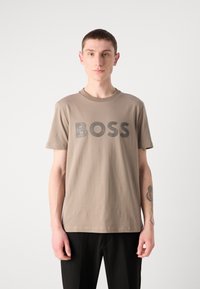 Šviesiai rudas marškinėliai su trumpomis rankovėmis ir apvaliu iškirpimu, ant priekinės dalies puikuojasi didelė juoda "BOSS" logotipas. Lygus paviršius.