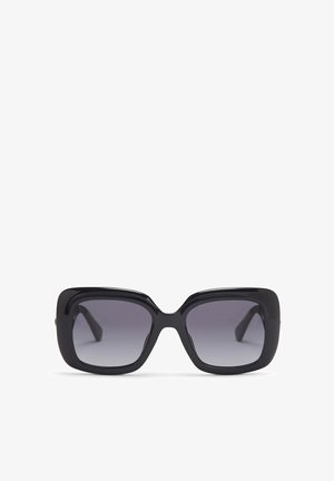 Lunettes de soleil noires oversize avec montures carrées, verres dégradés foncés et finition lisse et brillante. Elles présentent des branches épaisses avec des courbes subtiles.