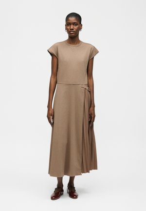 Kobieta stoi w bezrękawowej sukience midi w kolorze taupe z plisowanym bocznym panelem oraz brązowymi płaskimi butami na tle czystej białej ściany.