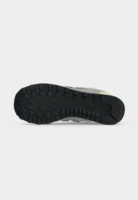 Semelle en caoutchouc noir pour basket avec des rainures profondes, logo visible de la marque près du talon et bords supérieurs de la chaussure gris clair.