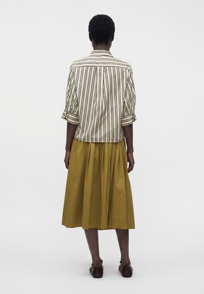 Personne aux cheveux courts portant une chemise rayée retroussée, une jupe midi plissée moutarde et des chaussures marron foncé, debout de dos.