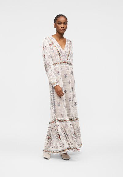 Femme portant une longue robe blanche avec une broderie florale et géométrique colorée, debout devant un fond clair uni.