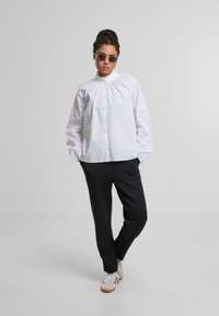 Weißes Hemd mit gekräuseltem Kragen, getragen mit schwarzen Hosen und weißen Sneakers. Das Model hat lockige Haare und trägt runde Sonnenbrillen.