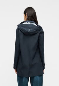 Γυναίκα που φοράει σκούρο ναυτικό αδιάβροχο με κουκούλα και μακριά μανίκια, φαίνεται από πίσω, συ νδυασμένο με χαλαρά μπλε τζιν σε απλό φόντο.