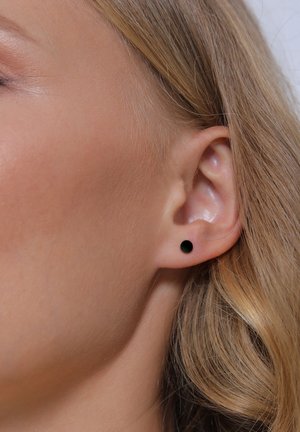 Gros plan sur l'oreille d'une femme aux cheveux blonds clairs et raides, portant une petite boucle d'oreille noire ronde.