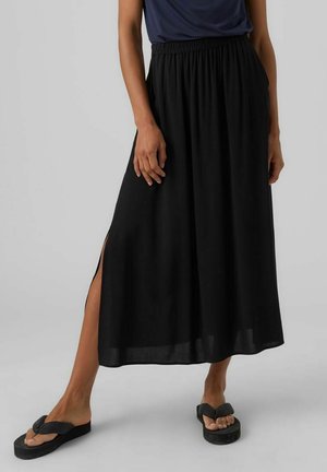 Schwarzer Maxi-Rock mit elastischem Bund, der seitliche Schlitze für mehr Bewegungsfreiheit aufweist. Getragen mit schwarzen Flip-Flops vor einfarbigem Hintergrund.
