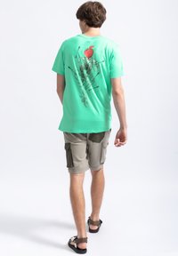 Mintgroene t-shirt met korte mouwen en een grafische print op de achterkant, waarop een figuur met een appelhoofd en pijlen staat afgebeeld, gecombineerd met beige cargoshorts.