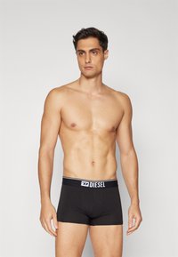 Μαύρο βαμβακερό σλιπ με λευκή ελαστική μέση που φέρει το λογότυπο "DIESEL". Λείος υφασμάτινος χαρακτήρας και εφαρμοστή σχεδίαση. Το μοντέλο στέκεται μπροστά.