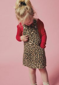 Haljina od trapera s uzorkom leoparda, s naramenicama, prednjim džepovima i detaljima od dugmadi, u kombinaciji s crvenim majicom dugih rukava i teksturiranim čarapama do koljena.