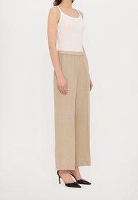 Pantalon large en lin beige avec taille élastique, associé à un débardeur blanc moulant et des talons noirs à bout pointu.