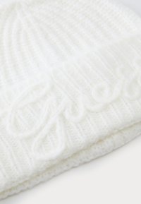 Un morbido berretto a maglia bianco con un motivo testurizzato e una scritta ricamata sul risvolto, poggiato su una superficie chiara.