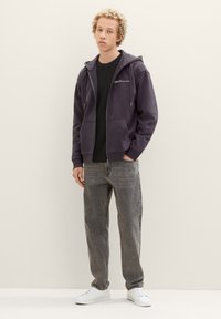 Lila Hoodie mit Reißverschluss und zwei Fronttaschen, getragen über einem schwarzen strukturierten Pullover, kombiniert mit hellgrauen Jeans und weißen Sneakers.