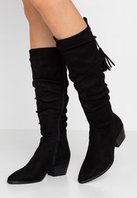 Bottes montantes en daim noir avec un design décontracté, bout pointu, talon effilé et un pompon décoratif sur le revers. Fermeture éclair latérale.