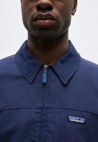 Κοντινό πλάνο ενός ατόμου που φοράει ένα σακάκι Patagonia σε ναυτικό μπλε με φερμουάρ στον γιακά, εστιάζοντας στο στήθος και την κάτω περιοχή του προσώπου.