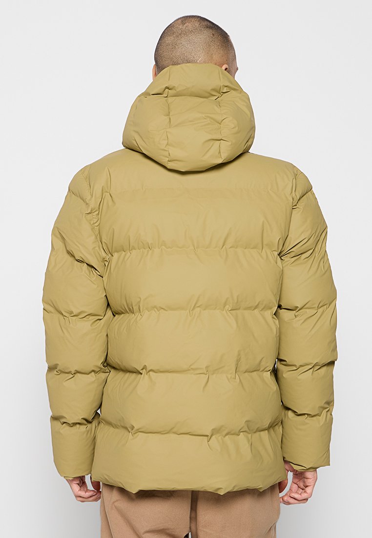 Man in een kaki pufferjack met capuchon, gezien vanuit de rug tegen een effen lichte achtergrond.
