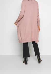 Lang roze cardigan van zacht stof, met lange mouwen, openingen aan de zijkanten en twee zijzakken, gecombineerd met zwarte broek en laarzen.